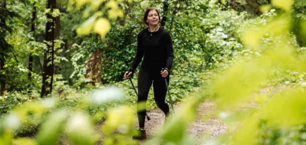 A woman hiking with poles on a forest trail surrounded by lush greenery.