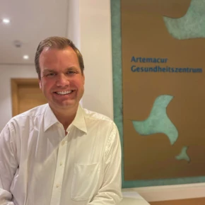 Ein lächelnder Mann in einem weißen Hemd steht an einem Empfangstresen im Artemacur Gesundheitszentrum.