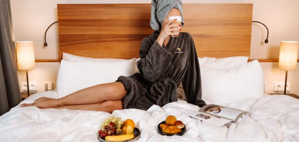 Eine entspannte Frau in einem Bademantel genie&szlig;t Fr&uuml;hst&uuml;ck im Bett, umgeben von gem&uuml;tlicher Atmosph&auml;re im Hotel Klosterhof.