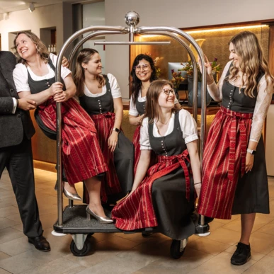 Group of people in traditional attire having fun with a luggage cart in Hotel Klosterhof's lobby.