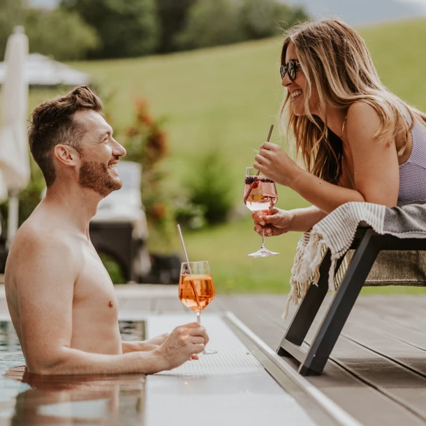 A couple enjoying drinks by the pool, with one person in the water and the other lounging on a chair, surrounded by a serene landscape.