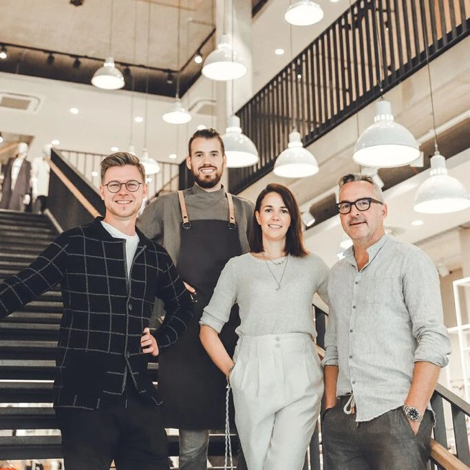 Eine Gruppe von vier Personen posiert lächelnd auf einer Treppe in einem modernen Gebäude mit dekorativen Lampen.