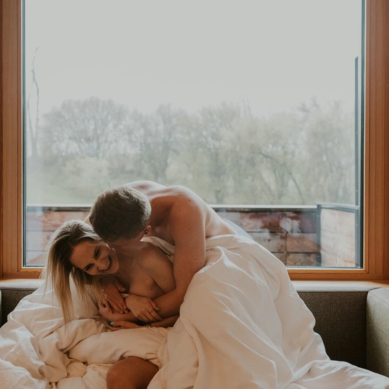 A couple laughs and embraces under a blanket in front of a large window with a nature view.