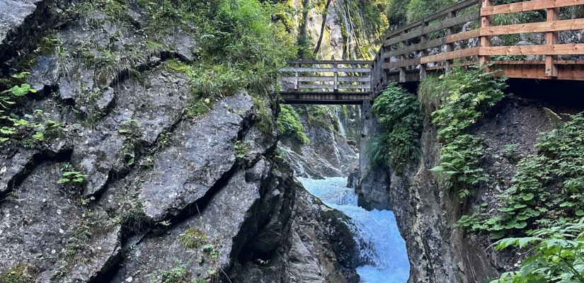 Ein malerischer Wanderweg mit einer Holzbrücke über einen klaren Bach, umgeben von grünen Pflanzen und steilen Felsen.