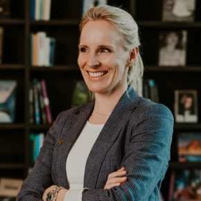 A smiling woman in a suit stands in front of a bookshelf.