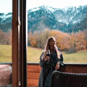 Frau in einem Bademantel genießt ein Glas Wein auf dem Balkon mit Blick auf die Berge und die herbstliche Landschaft.
