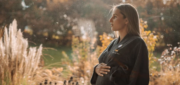 Eine schwangere Frau in einem Bademantel steht in der Natur, umgeben von herbstlichen Pflanzen, im Hotel Klosterhof, Bayern.