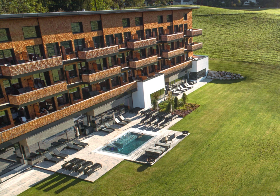 Moderne Hotelansicht mit Terrasse und Liegen, umgeben von gepflegtem Grün und einem kleinen Pool im Vordergrund.