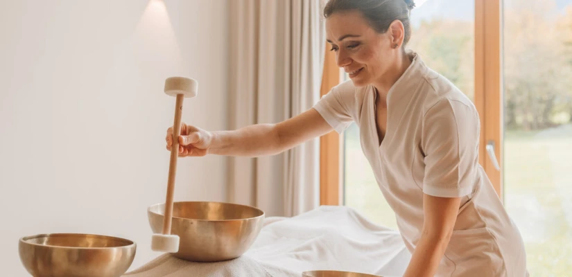 A woman performs a sound therapy session with singing bowls in a tranquil spa setting.