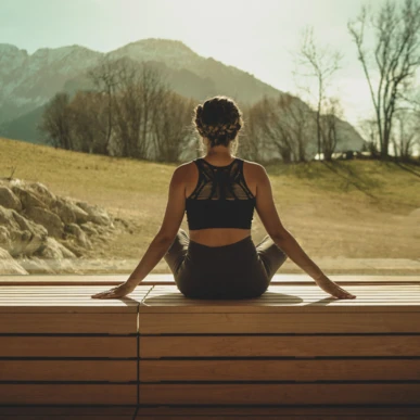 A person in a yoga pose sits inside, gazing at the serene mountain view through large windows at Wellnesshotel Klosterhof in Bayern.