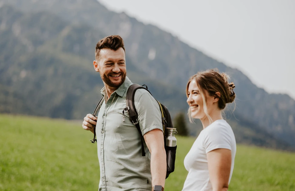 Ein Paar wandert lächelnd in der Nähe des Hotel Klosterhof in Bayerisch Gmain, mit grünen Wiesen und Bergen im Hintergrund.