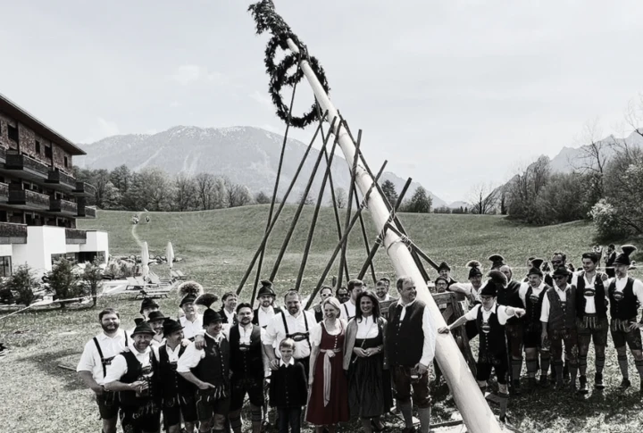 Eine Gruppe in traditioneller Tracht stellt einen Maibaum auf einer Wiese vor dem Hotel Klosterhof auf, im Hintergrund Berge.