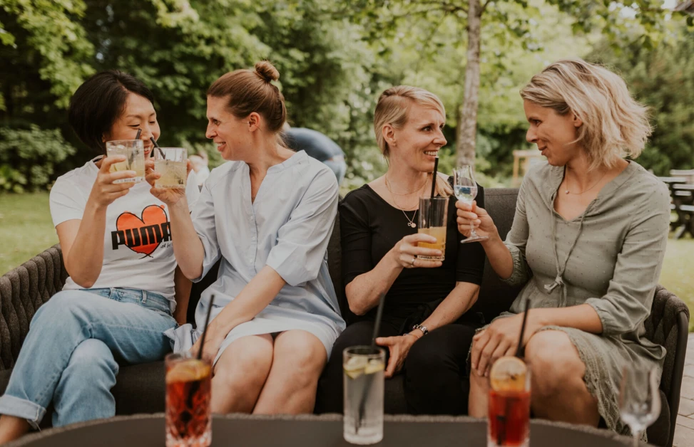 Fünf Frauen genießen Drinks auf einer Couch im Freien, umgeben von grünem Ambiente. Freundliche Stimmung und geselliges Beisammensein.