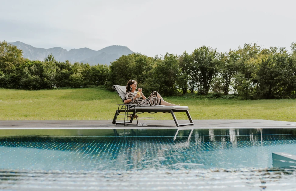 Eine Frau entspannt auf einer Liege am Pool, umgeben von grünen Wiesen und Bergen im Hintergrund.