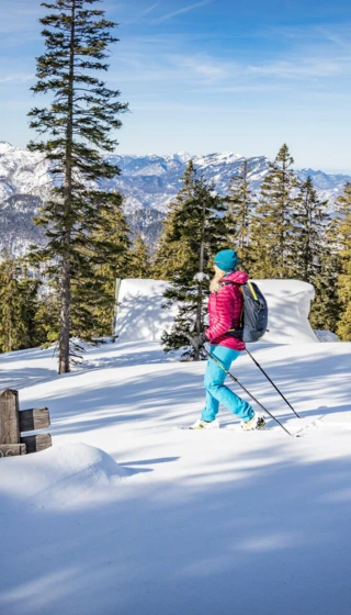 Eine Person wandert durch verschneite Landschaften mit Tannenbäumen, in der Nähe eines Wegweisers, mit Bergen im Hintergrund.