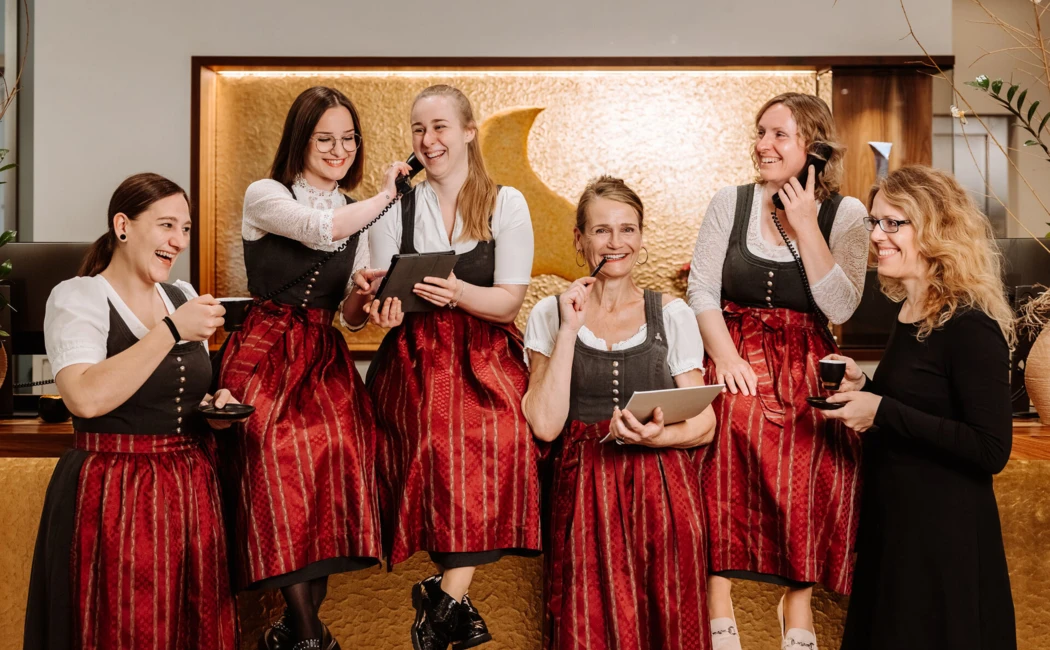 Sechs Frauen in traditioneller Tracht lachen und arbeiten in einem modernen Empfangsbereich. Einladende Atmosphäre und Teamgeist.
