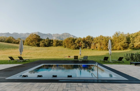 Ein moderner Pool umgeben von Liegen und Sonnenschirmen, mit Blick auf eine grüne Wiese und die Berge im Hintergrund.