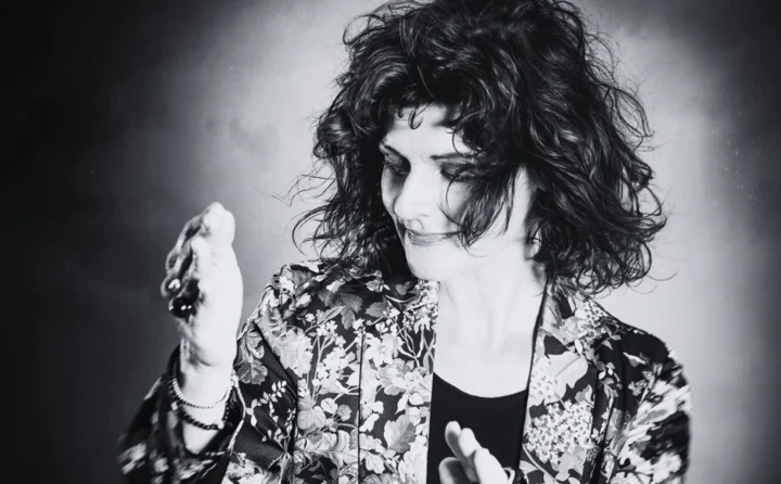 Eine Frau mit lockigem Haar in einem blumigen Blazer, die mit einer Handbewegung in einem künstlerischen Moment posiert.