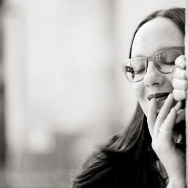 Eine Frau mit Brille lächelt sanft und lehnt sich an eine Wand, in einem entspannten Moment.