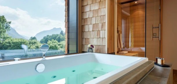 Blick auf eine Badewanne mit grünem Wasser und Aussicht auf Berge durch große Fenster in einem modernen Wellnessbereich.