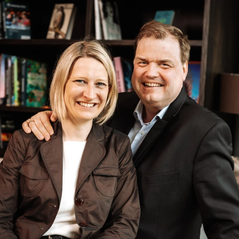 Ein lächelndes Paar posiert entspannt in einem modernen Innenraum des Hotel Klosterhof, umgeben von Bücherregalen.