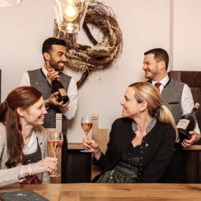 People in traditional attire enjoying wine at a wooden table, smiling and toasting.