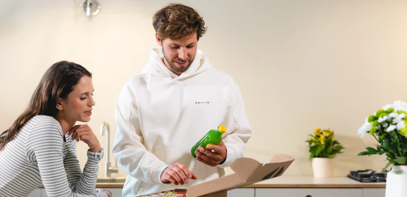 Junger Mann öffnet eine Box in der Küche, während eine Frau interessiert zuschaut. Frische Getränke und Pflanzen im Hintergrund.