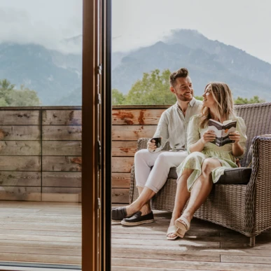 A couple relaxes on a terrace with mountain views, enjoying a book and coffee.