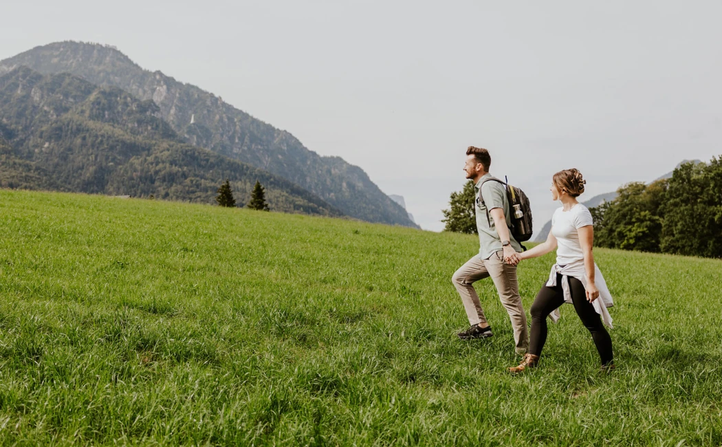 Ein Paar wandert Hand in Hand &uuml;ber eine gr&uuml;ne Wiese mit Bergen im Hintergrund.