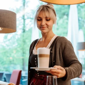 Eine Kellnerin serviert einen Latte Macchiato in einem gemütlichen Café, umgeben von frischem Gebäck und einer einladenden Atmosphäre.