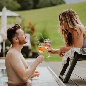 A man in a pool and a woman on a lounge chair toast with drinks, set against a scenic, lush background.