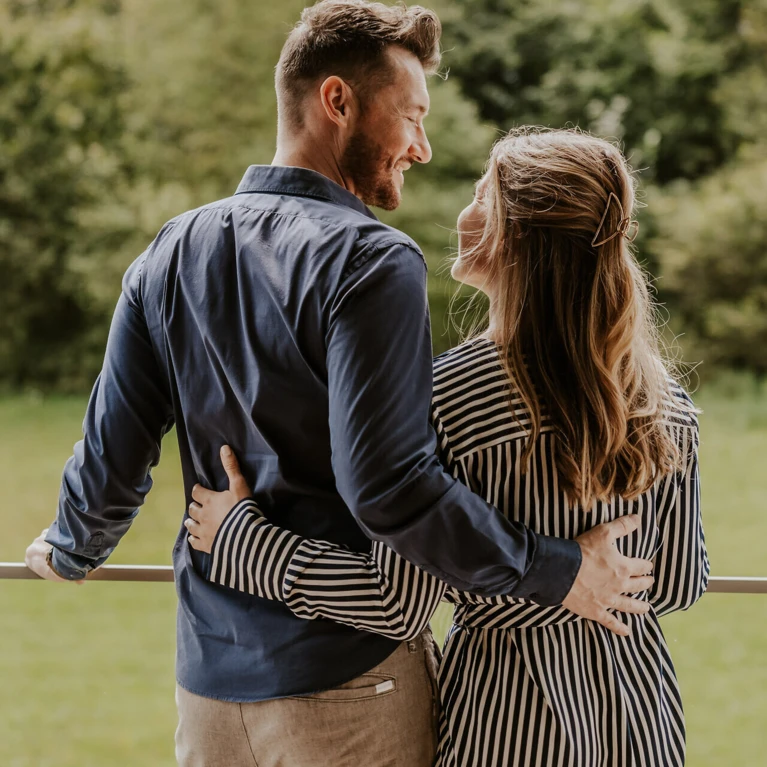 Ein Paar steht auf einem Balkon und blickt lächelnd in die Natur, umgeben von Bäumen und Bergen im Hintergrund.