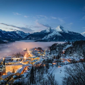 Blick auf eine beleuchtete Stadt in den verschneiten Alpen bei Dämmerung, umgeben von majestätischen Bergen und Nebel.