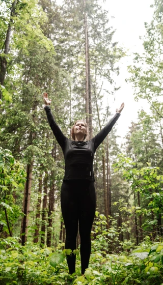 Eine Person steht in einem Wald mit erhobenen H&auml;nden, umgeben von gr&uuml;nen Pflanzen und hohen B&auml;umen, und genie&szlig;t die Natur.