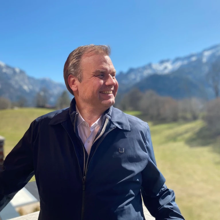 Ein Mann steht im Freien, lächelt und genießt die Aussicht auf eine grüne Wiese und schneebedeckte Berge im Hintergrund.