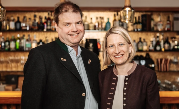 Ein Paar lächelt vor einer Bar mit einer Auswahl an Getränken im Hintergrund, beide tragen traditionelle Kleidung.