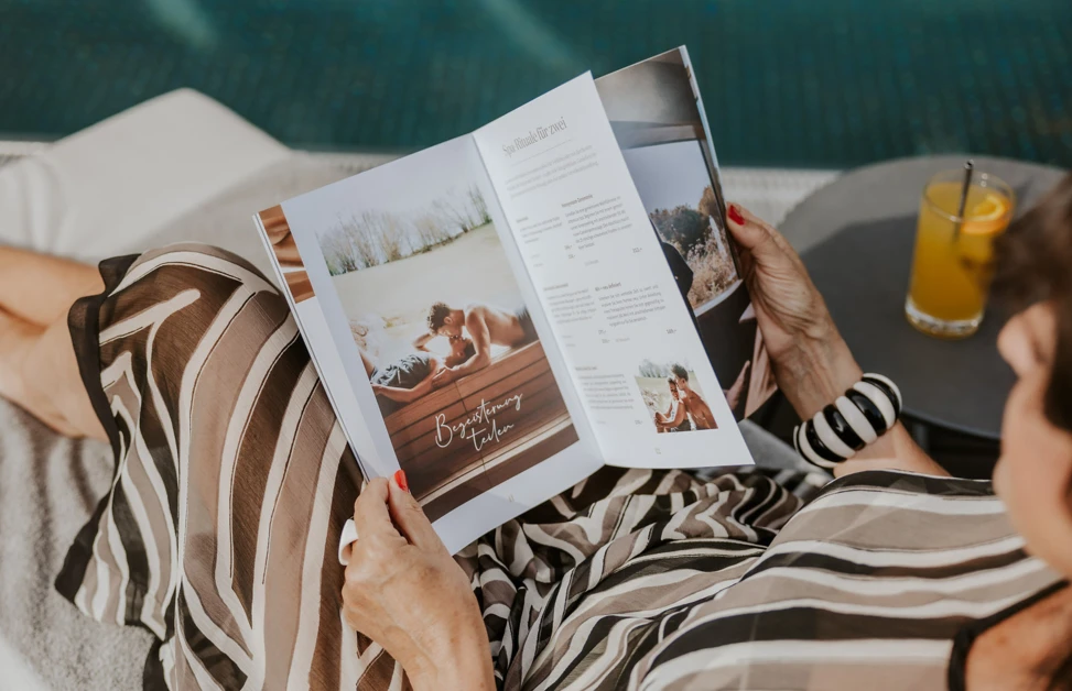 Eine Person liest eine Brosch&uuml;re am Pool, mit einem Getr&auml;nk auf dem Tisch.