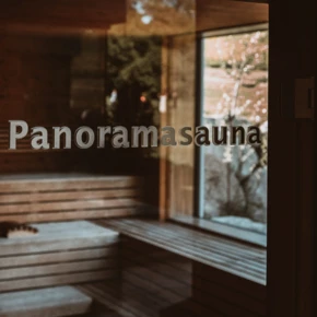 Glasierte Tür mit dem Schriftzug "Panoramasauna", Blick auf die Holzbank im Innenraum des Wellnessbereichs.
