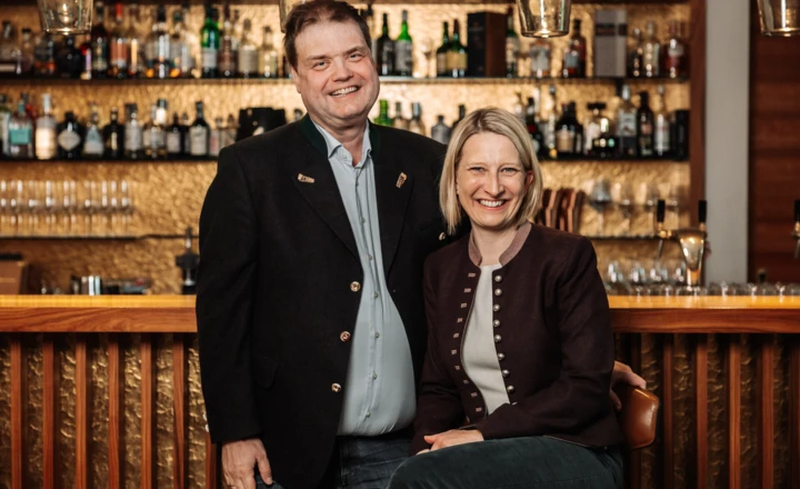 A smiling man and woman stand together in front of a well-stocked bar, exuding warmth and hospitality.