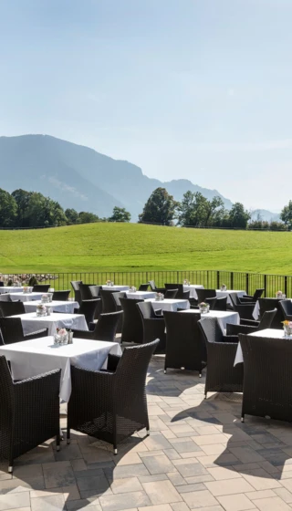 Sonnige Terrasse mit Blick auf die Berge, elegante Tische und Stühle, umgeben von grüner Landschaft. Perfekt für entspannte Mahlzeiten.