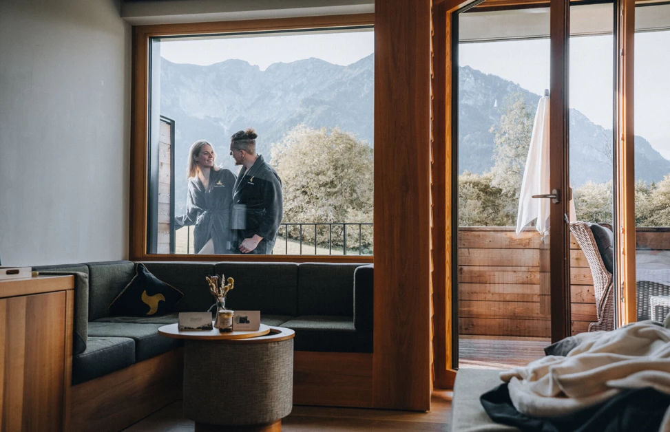 Ein Paar in Bademänteln steht an einem Fenster mit Blick auf die Berge, umgeben von einem modernen, gemütlichen Raum.