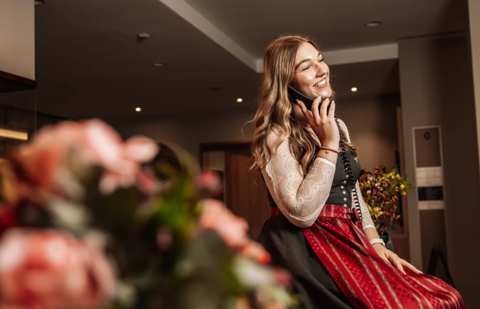 Eine lächelnde Frau im traditionellen Outfit telefoniert in der Lobby des Hotel Klosterhof, umgeben von Blumenarrangements.