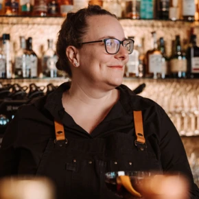 Eine Barkeeperin mit Brille hält ein Cocktailglas in der Hand, umgeben von einer Auswahl an Spirituosen im Hotel Klosterhof, Bayern.