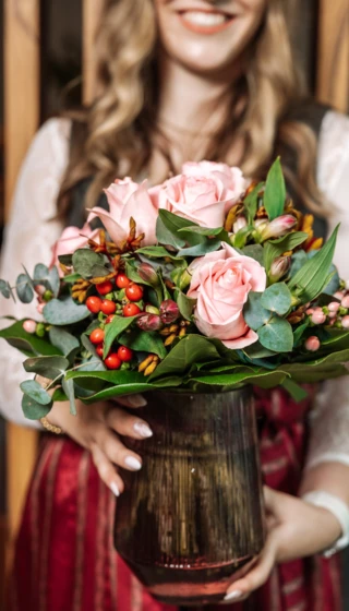 Eine Frau hält einen Strauß mit rosa Rosen und buntem Grün in einer Vase, lächelnd vor einem Holz-Hintergrund.
