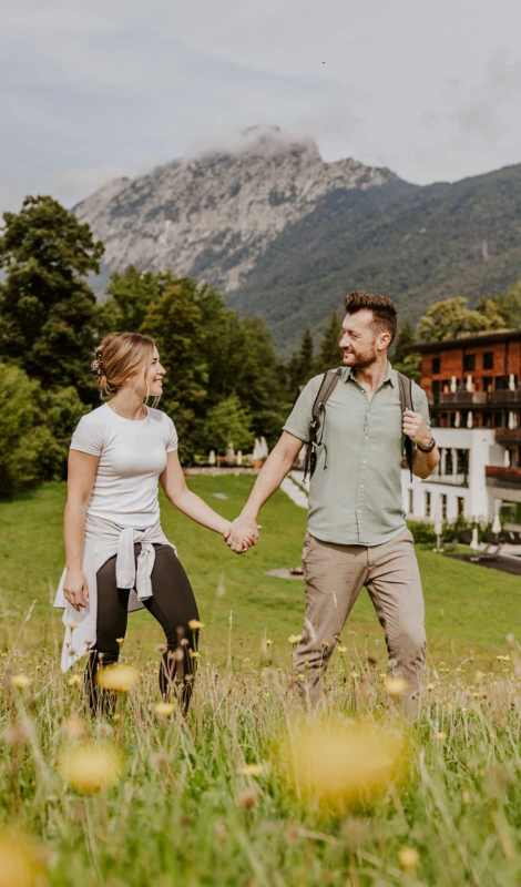 Ein Paar hält Händchen und spaziert durch eine blühende Wiese mit Bergen im Hintergrund, umgeben von Natur und frischer Luft.