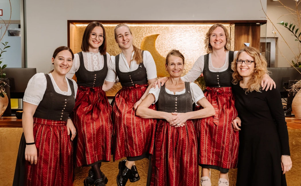 Sechs Frauen in traditioneller Tracht stehen lächelnd in der Lobby des Hotel Klosterhof, umgeben von modernen Elementen.