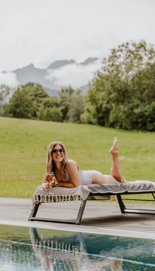 A woman lounges on a sunbed by a pool, holding a drink, with a scenic view of trees and mountains in the background.