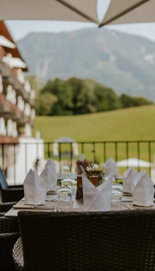 Outdoor dining at Wellnesshotel Klosterhof in Bayern, featuring neatly set tables with folded napkins and a view of lush green hills and mountains.