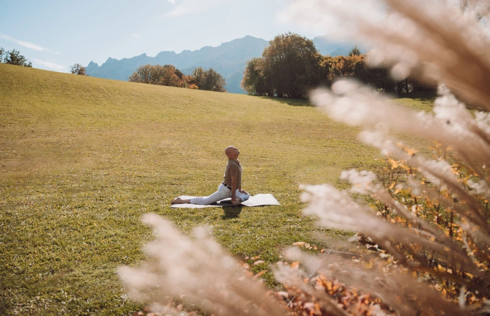 Eine Person meditiert auf einer Matte in einer grünen Wiese, umgeben von Bergen, ideal für Wellness und Entspannung im Hotel Klosterhof.