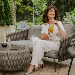 A woman in elegant attire smiles while holding a drink, seated outdoors on a stylish patio.